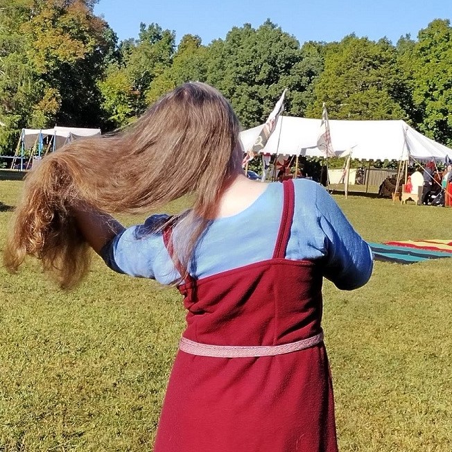 The back of a woman standing outside brushing her long brown hair, wearing a bright blue dress with a dark red apron over it