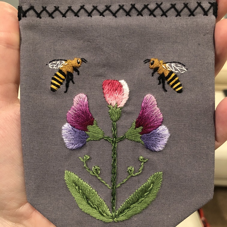 A grey cloth hand embroidered with two bees and three sweet pea flowers