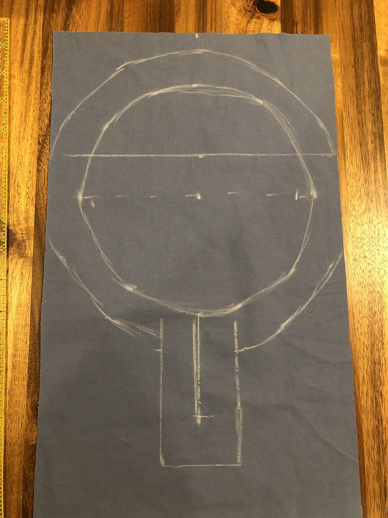 A blue piece of fabric with a keyhole neckline pattern drawn in white chalk. The lines of the pattern are crooked and messy