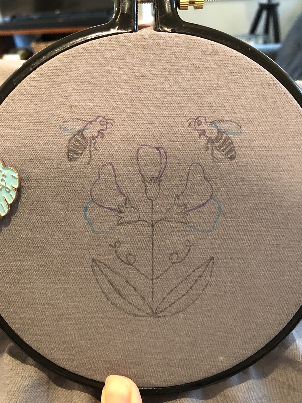 A grey cloth in an embroidery hoop with a drawing of two bees and three sweet pea flowers on the cloth