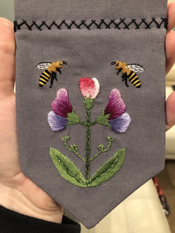 A grey cloth hand embroidered with two bees and three sweet pea flowers