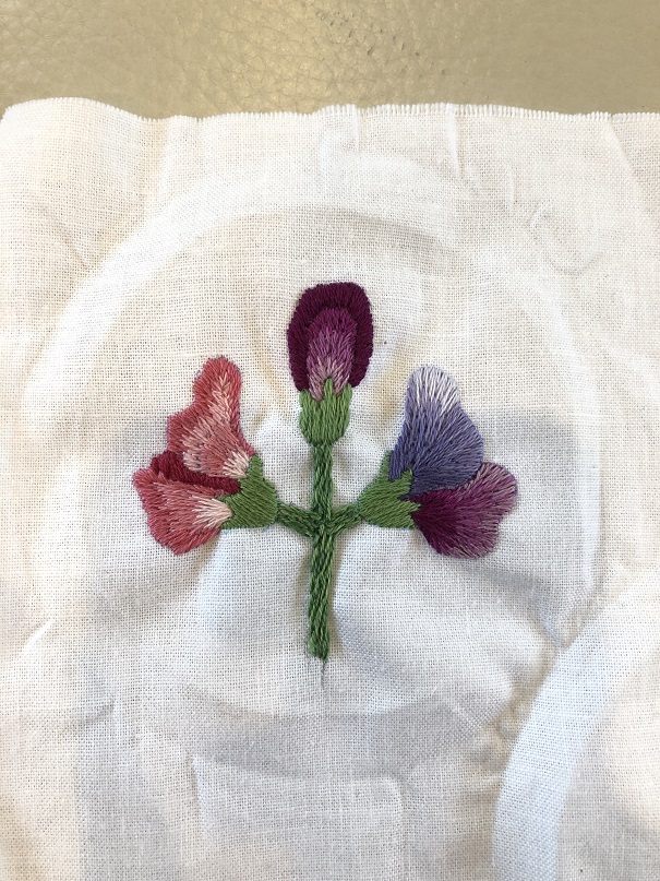 A piece of embroidery with three embroidered sweet pea flowers in pink and purple