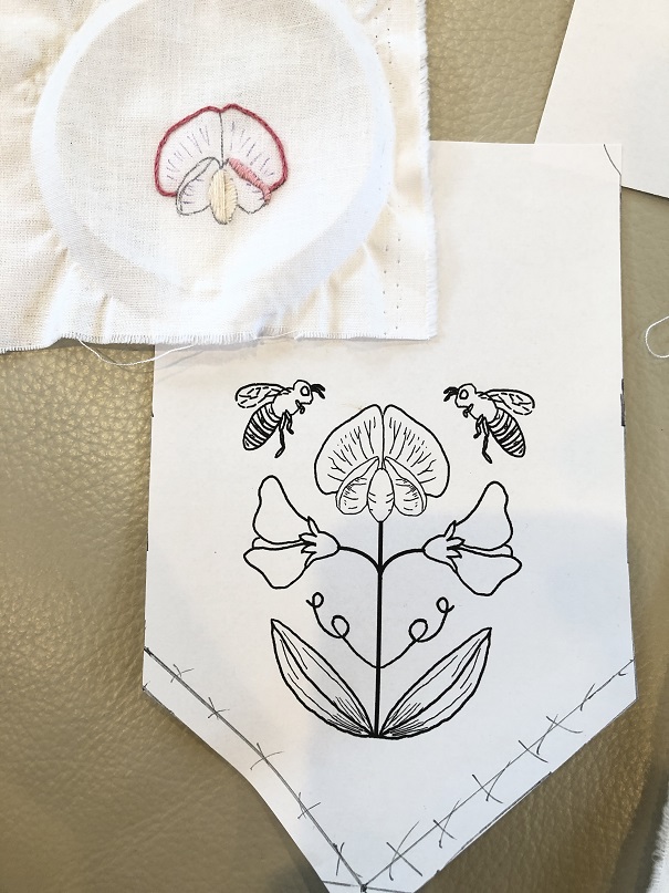 A piece of paper with a black and white printed design of three sweet peas different than the prior designs and two honey bees. A small incomplete embroidery of a sweet pea flower is on top of the paper.