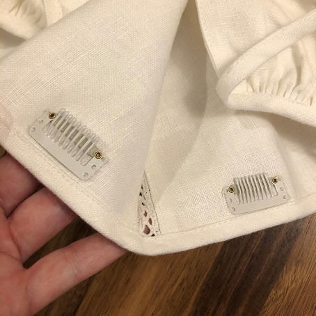 A close up of two white wig clips sewn into the front edge of a white linen coif