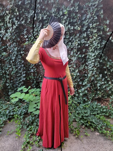 A woman wearing a red linen dress with gold silk sleeves pinned on. Her head is covered by a white veil, and she is also wearing a red, pearl and brass necklace as well as a black belt and brown shoes