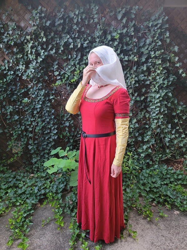 A woman wearing a red linen dress with gold silk sleeves pinned on. Her head is covered by a white veil, and she is also wearing a red, pearl and brass necklace as well as a black belt and brown shoes