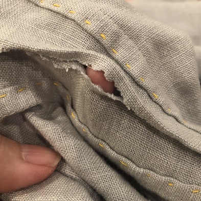 A photo of a finger poking through a small hole in the seam of a light grey tunic