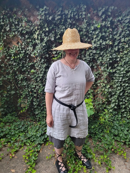 A photo of a woman standing in front of ivy wearing a straw hat that covers her face, a light grey tunic, dark grey shorts, a black belt, and black gilly shoes