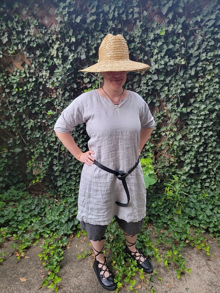 A second photo of a woman standing in front of ivy wearing a straw hat that covers her face, a light grey tunic, dark grey shorts, a black belt, and black gilly shoes