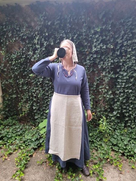 A woman in a dark blue dress wearing a natural linen apron that falls from her waist to the hem of her dress