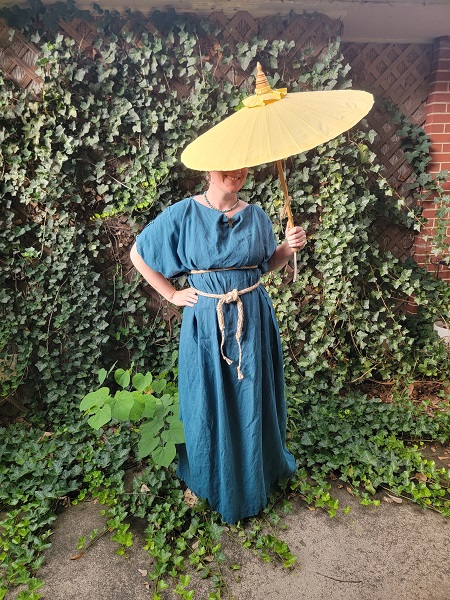 A woman wearing a teal gap-sleeve chiton, belted under the bust and at the waist witha rope belt, standing in front of ivy