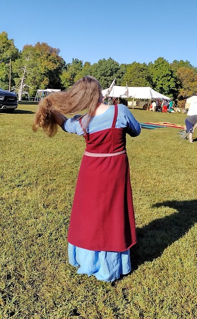 The back of a woman standing outside brushing her long brown hair, wearing a bright blue dress with a dark red apron over it