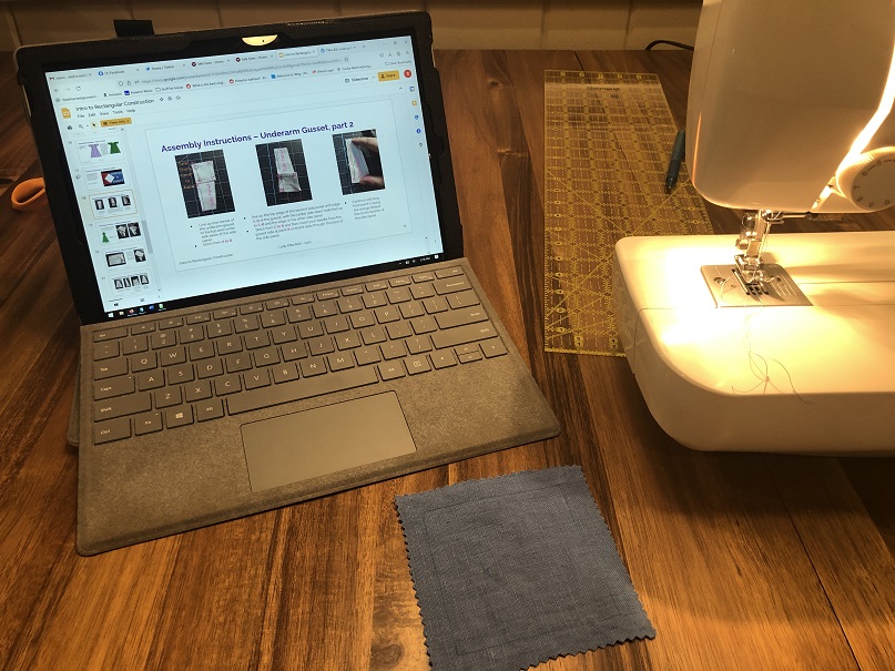 A photo of a wooden table with a laptop, a sewing machine, and a blue square of fabric. The screen on the laptop shows a slide from a presentation describing how to attach the blue square to the trapezoidal side panels