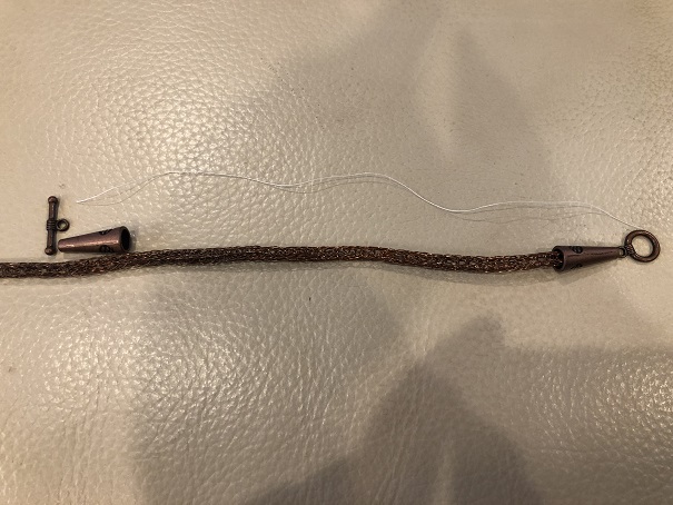 A length of woven copper wire laid out on a white leather footstool to estimate the length of the finished bracelet, with unattached end caps at each end and an unattached circle clasp