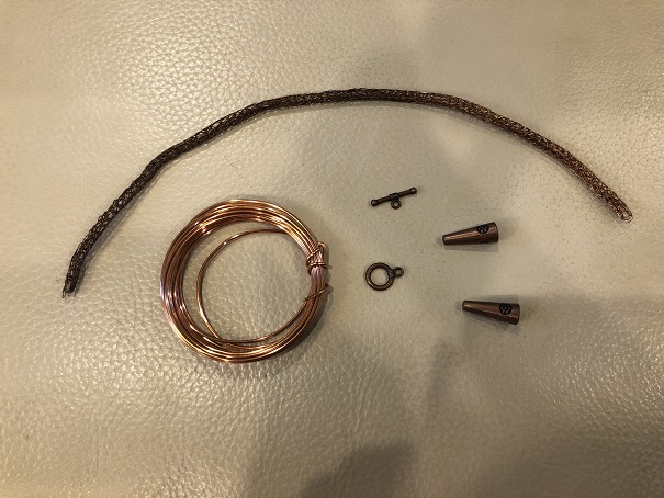 Several items arranged on a white leather footstool. Those items are a length of copper woven wire, a coil of thicker wire, two end caps, and a circle clasp set.