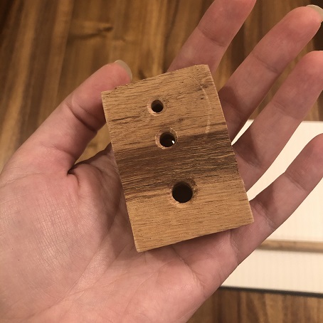 A hand holding a block of wood with three holes of different sizes drilled into it