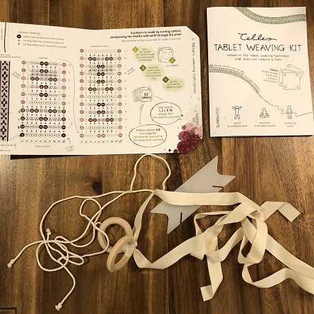 A photo showing a set of instructions for tablet weaving, a plastic shuttle, and a set of ribbons attached to a ring