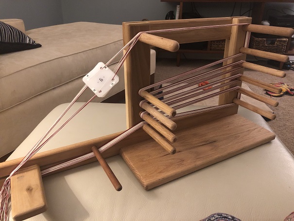 A photo of a wooden inkle loom with red and white thread woven around the pegs