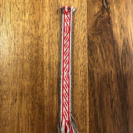 A close up of a piece of red, light grey, and dark grey tablet woven trim with a pattern that looks like angled box shapes