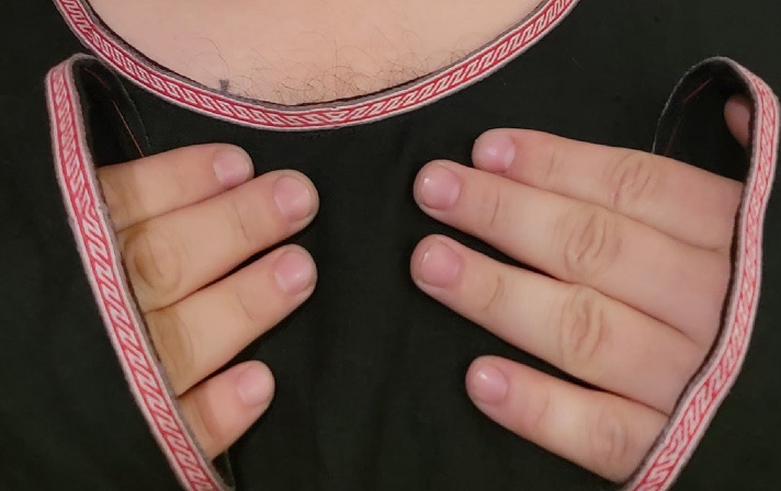 A photo showing a man wearing a black tunic with red, light grey, and dark grey tablet woven trim attached to the neckline and wrist cuffs of the tunic