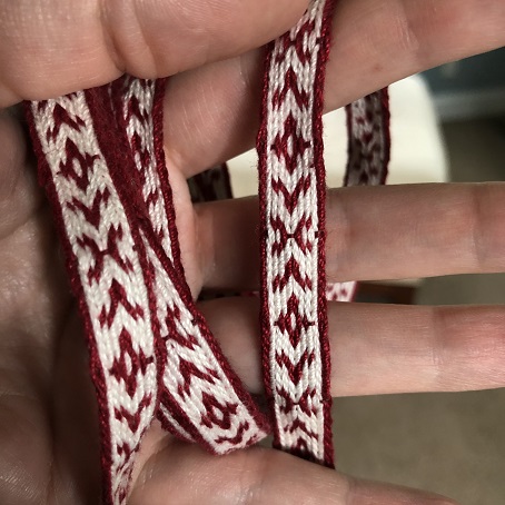 A close up of dark red and white tablet woven trim with a pattern of crosses and arrows