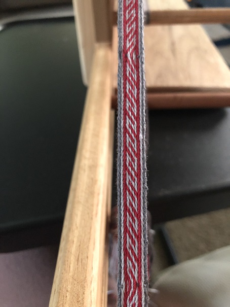 A photo of red, light grey, and dark grey tablet woven trim currently still on the inkle loom, with a pattern that looks like connected S and Z shapes